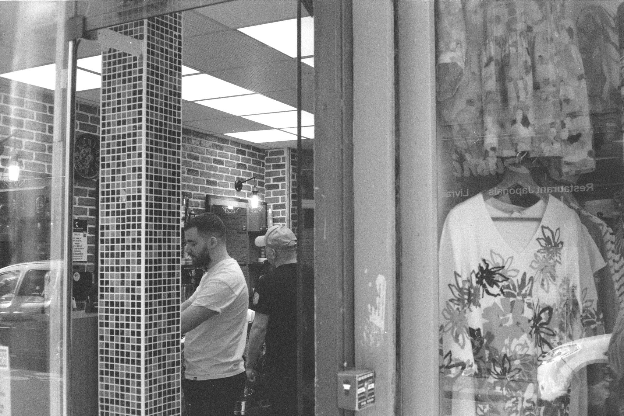 Two barbers at work behind the open glass front of a barbershop, beside a dress shop window