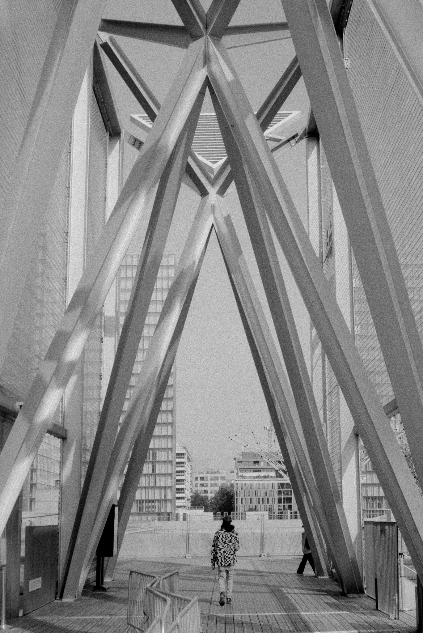 A lone walker passing beneath the massive crossed steel beams of a modern building
