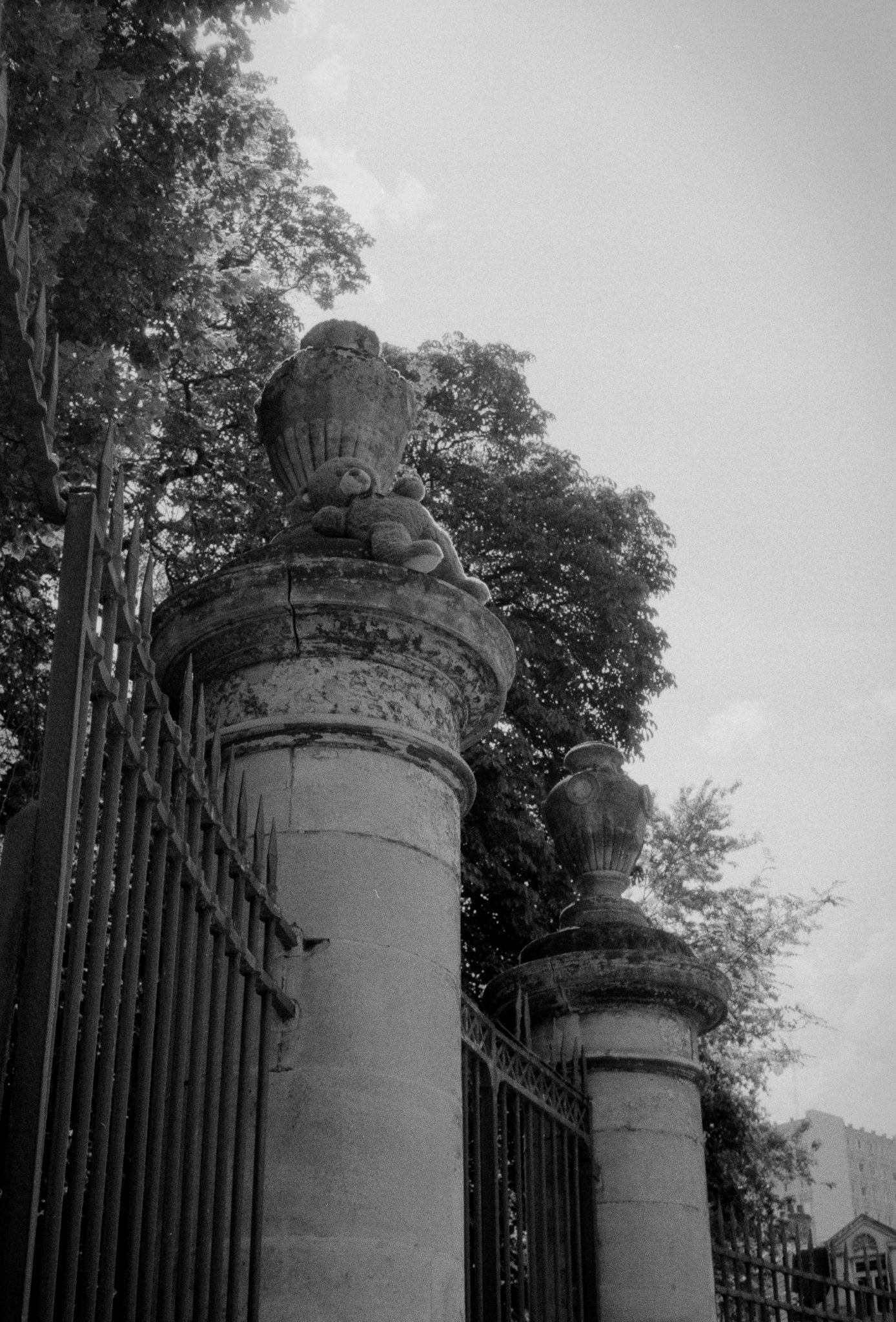 A worn teddy bear resting against a stone urn on top of a spiked iron gate pillar
