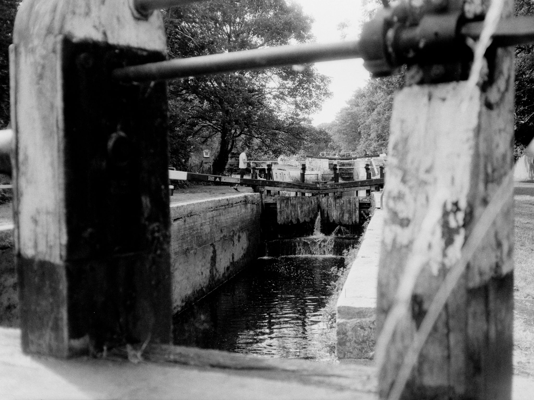 A canal lock framed by weathered wooden gate beams, water spilling through as a walker passes behind