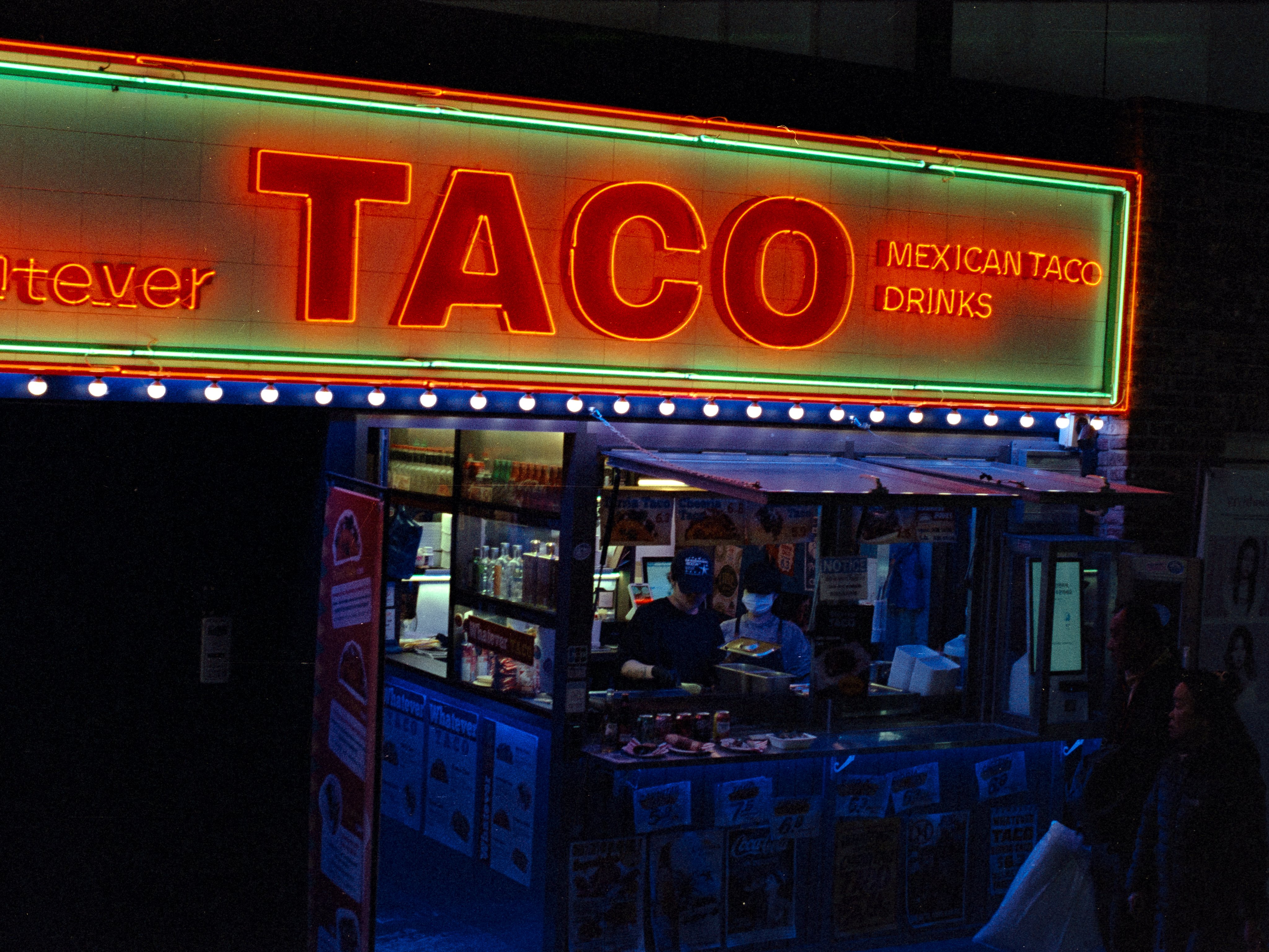 A neon-lit taco stand at night, two cooks working under the glowing red sign
