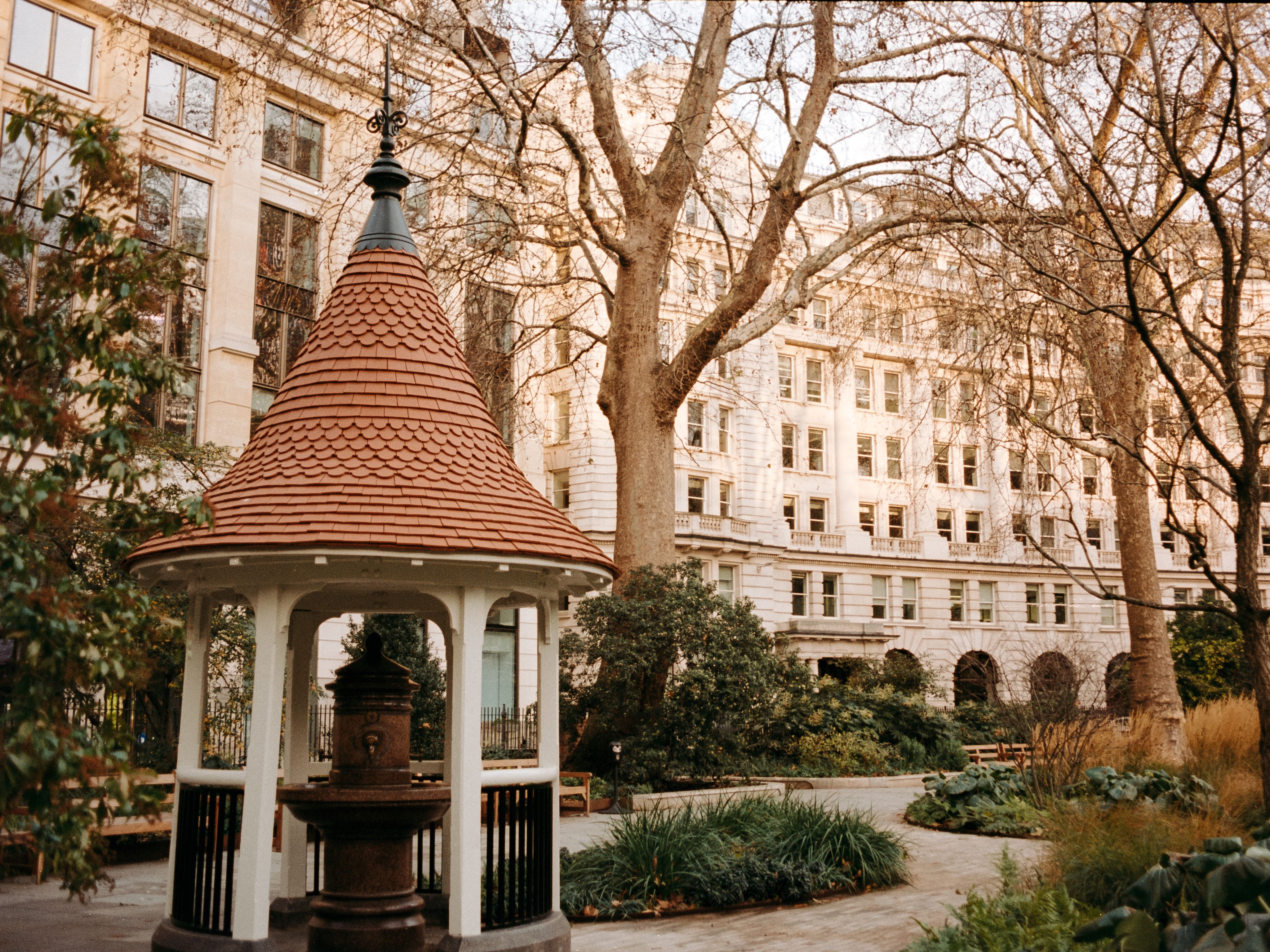 A gazebo with a conical tiled roof sheltering an old drinking fountain in a leafy city garden