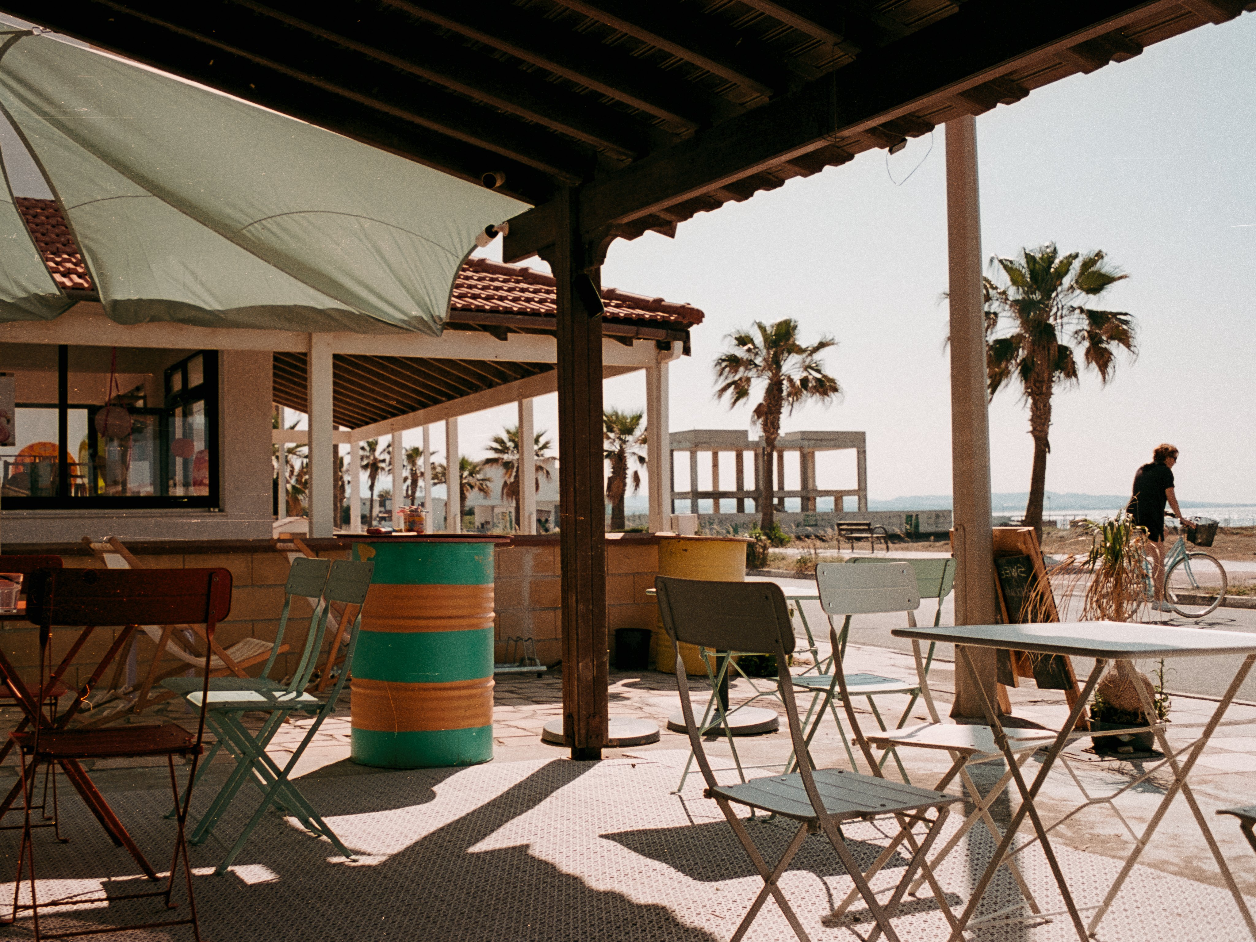 A beach bar terrace with pastel folding chairs and oil drum tables, palm trees and a concrete ruin behind