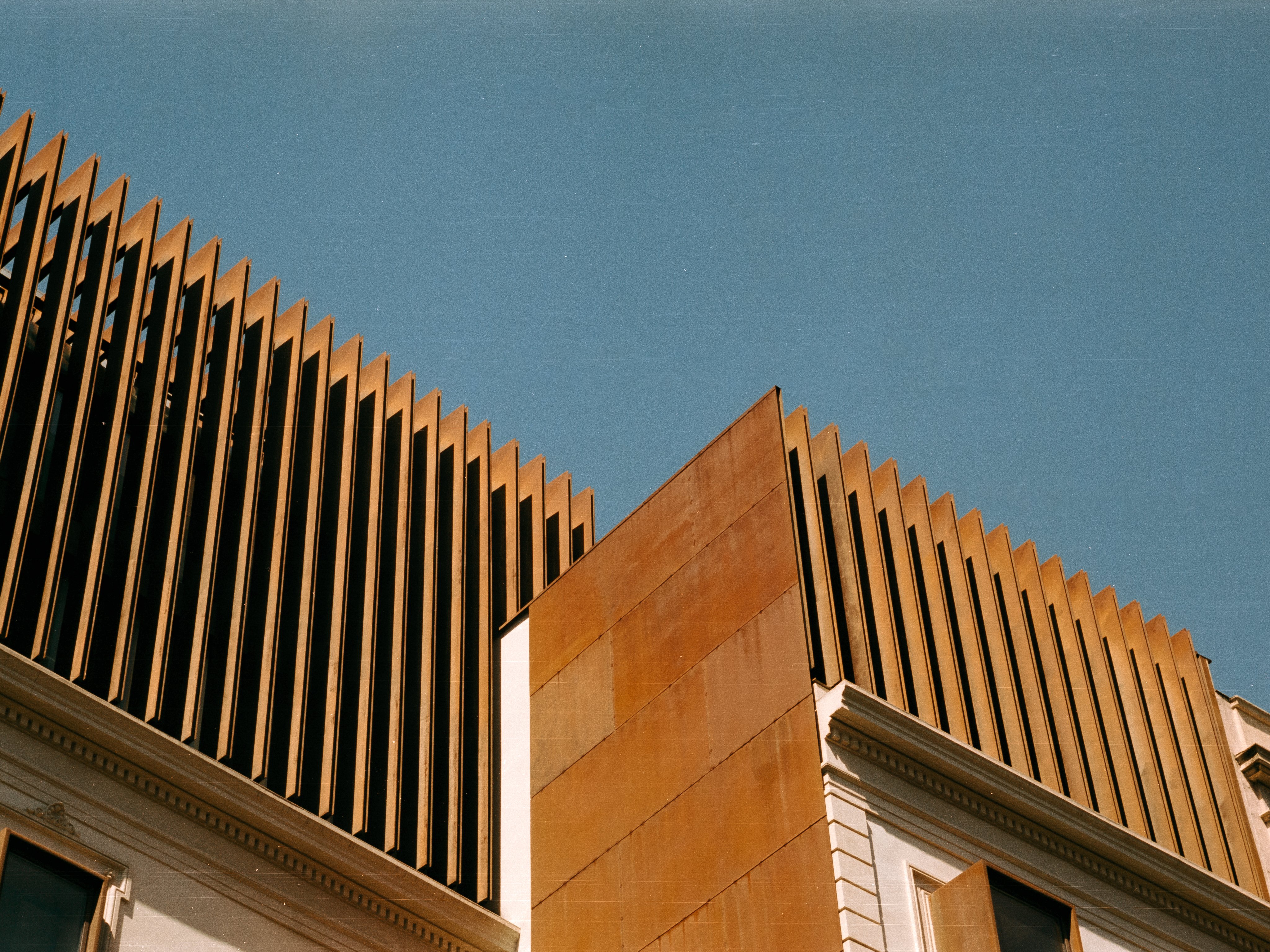 Rows of rusted steel fins crowning a classical facade against a clear blue sky